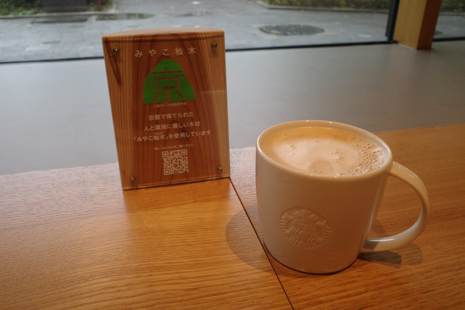 みやこ杣木のロゴが入った木のサインと、カウンターに置かれたスターバックスのマグカップ。窓の外は雨で濡れた敷石