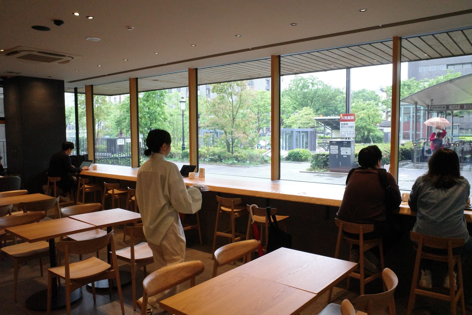 市役所広場側の長いカウンター席と手前のテーブル席。広場の緑と本庁舎の入口、雨の中を歩く人の姿が窓の外に広がる