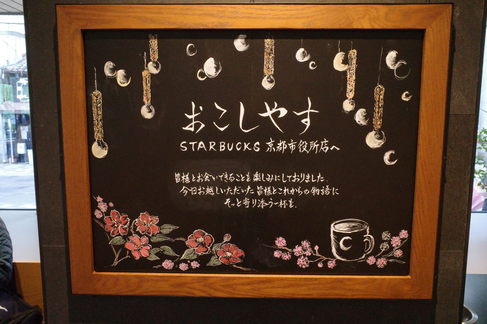 「おこしやす STARBUCKS 京都市役所店へ」と白チョークで書かれた黒いウェルカムボード。提灯と椿の花のイラスト