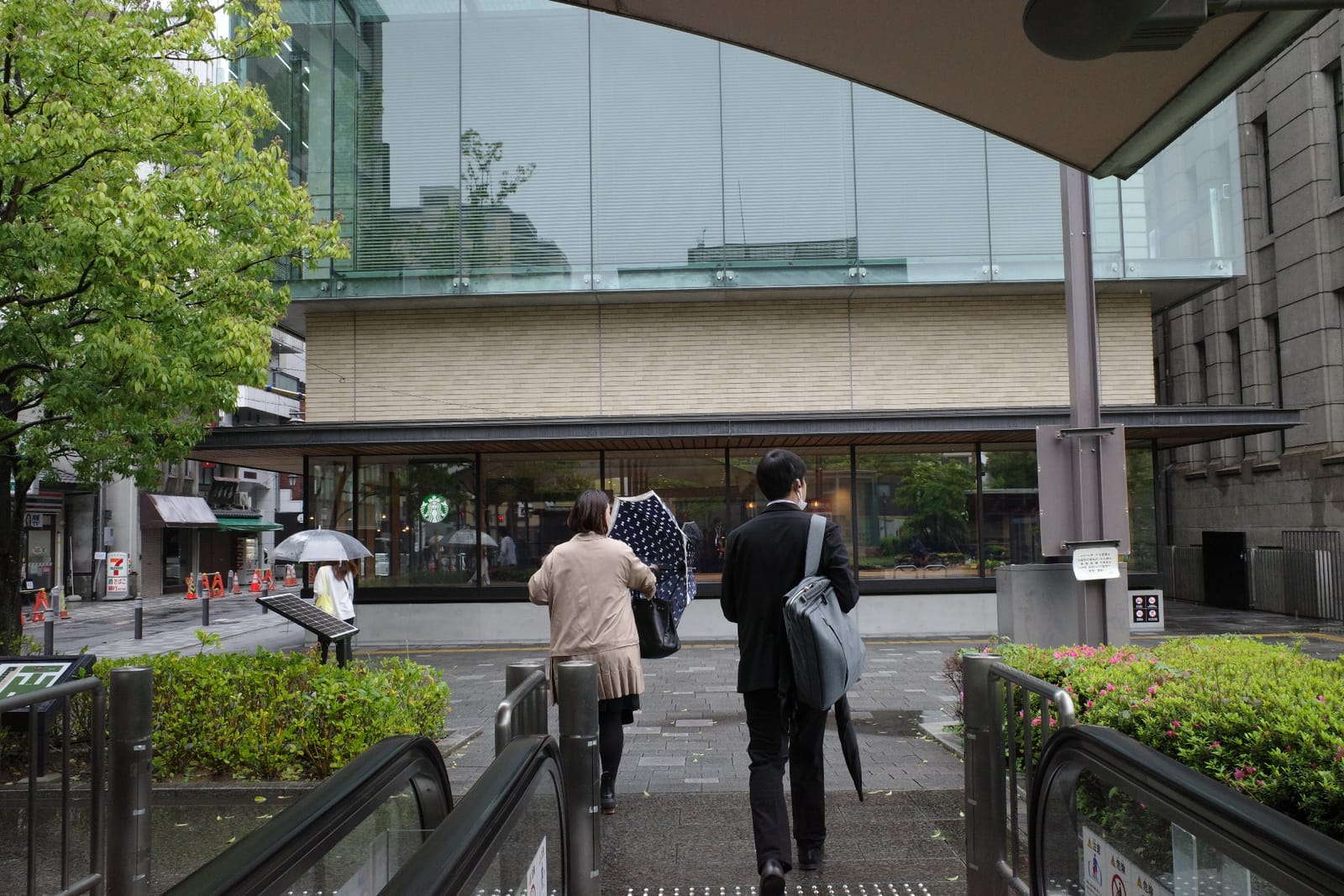Zestの出口を上がった先、雨に濡れた敷石越しに見える京都市役所店の大きなガラス窓と木のファサード